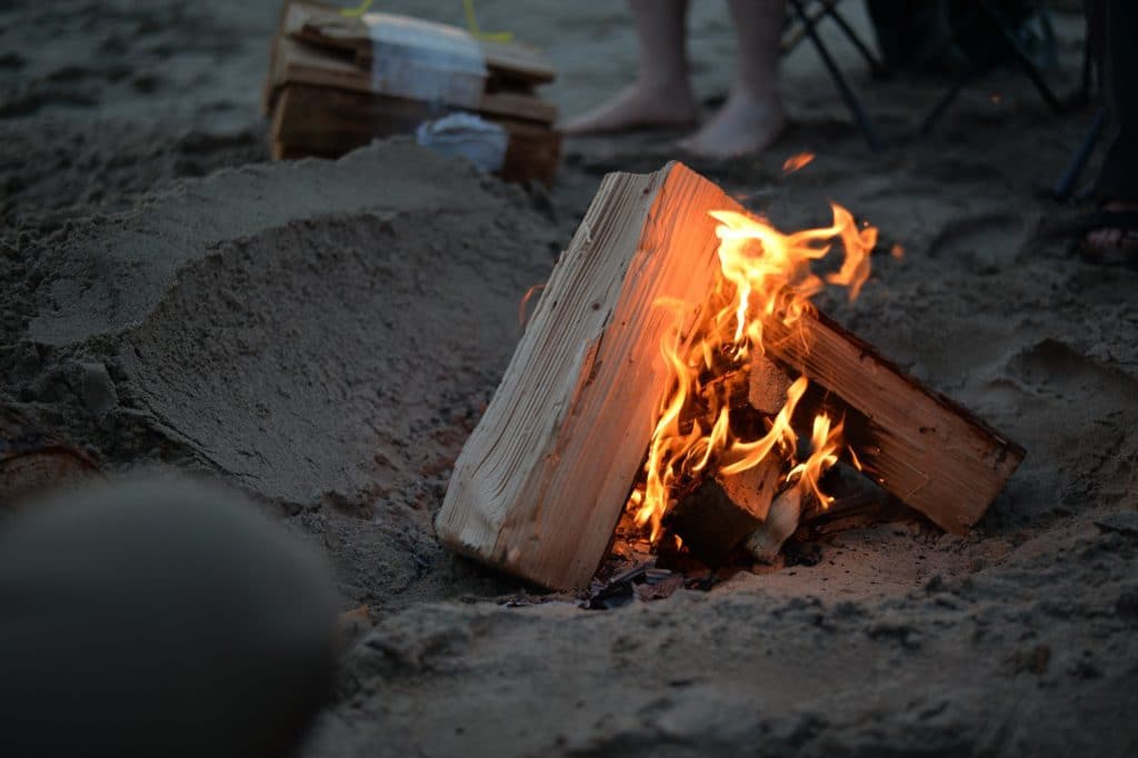 an image of fire wood burning on the beach at sunset with a camping chair and feet in the background - 11 Fun Things To Do On This Labor Day Weekend
