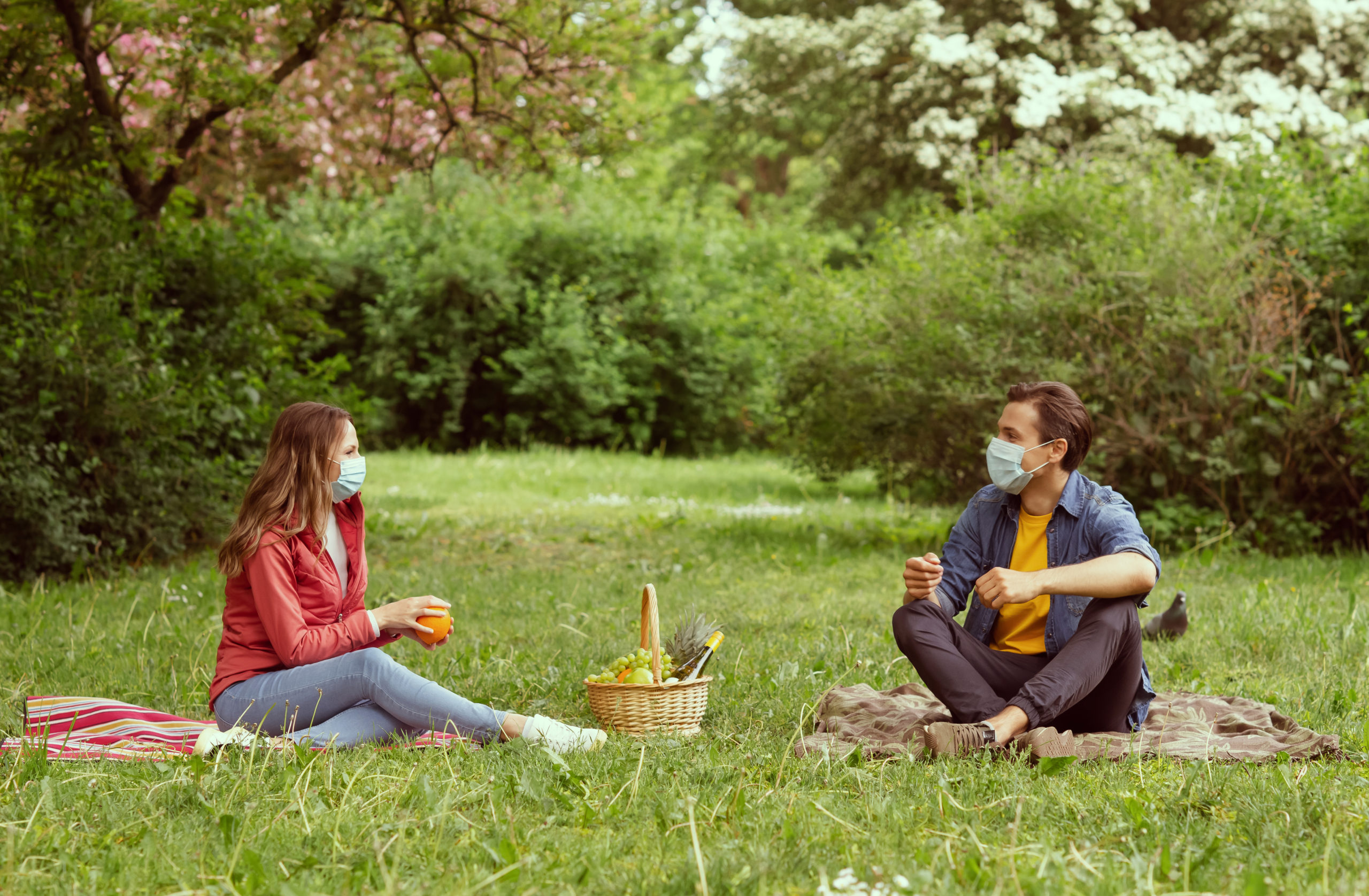 two people sitting in a park having a picnic practicing social distancing and wearing face masks
